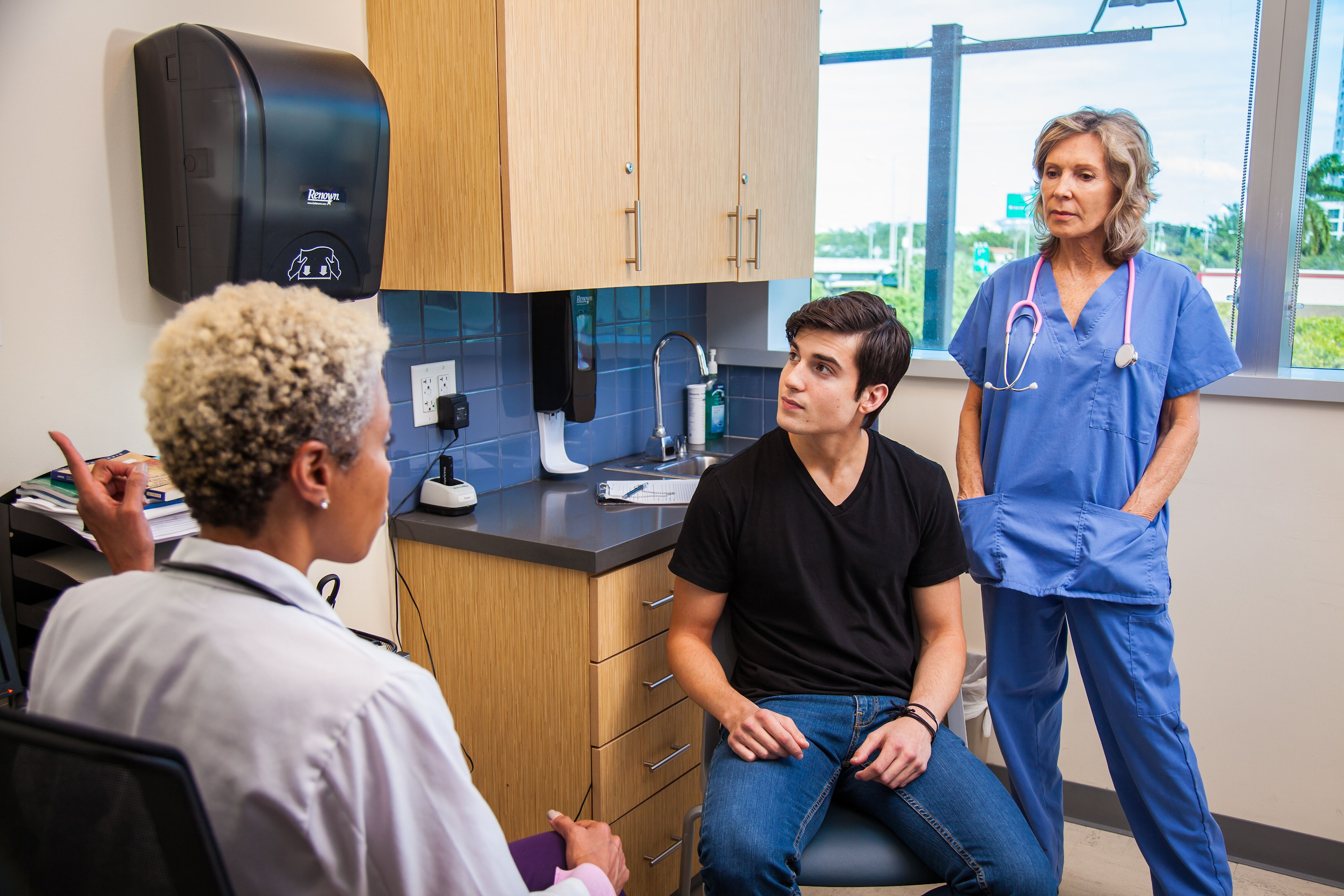 A seated clinician speaks to a seated client while a medical interpreter stands behind the client.